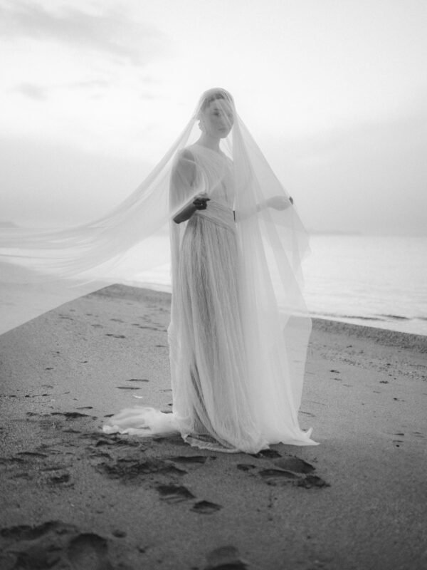 Destination Elopement in Rethymno, Crete, black and white image of a bride with veil on, standing on the beach looking like Madonna
