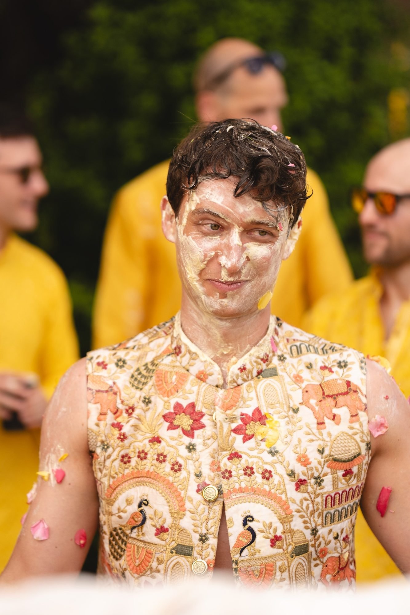 Groom in blazer with Indian motif smiling while being painted at beachfront Haldi and Mehndi celebration in the Athenian Riviera