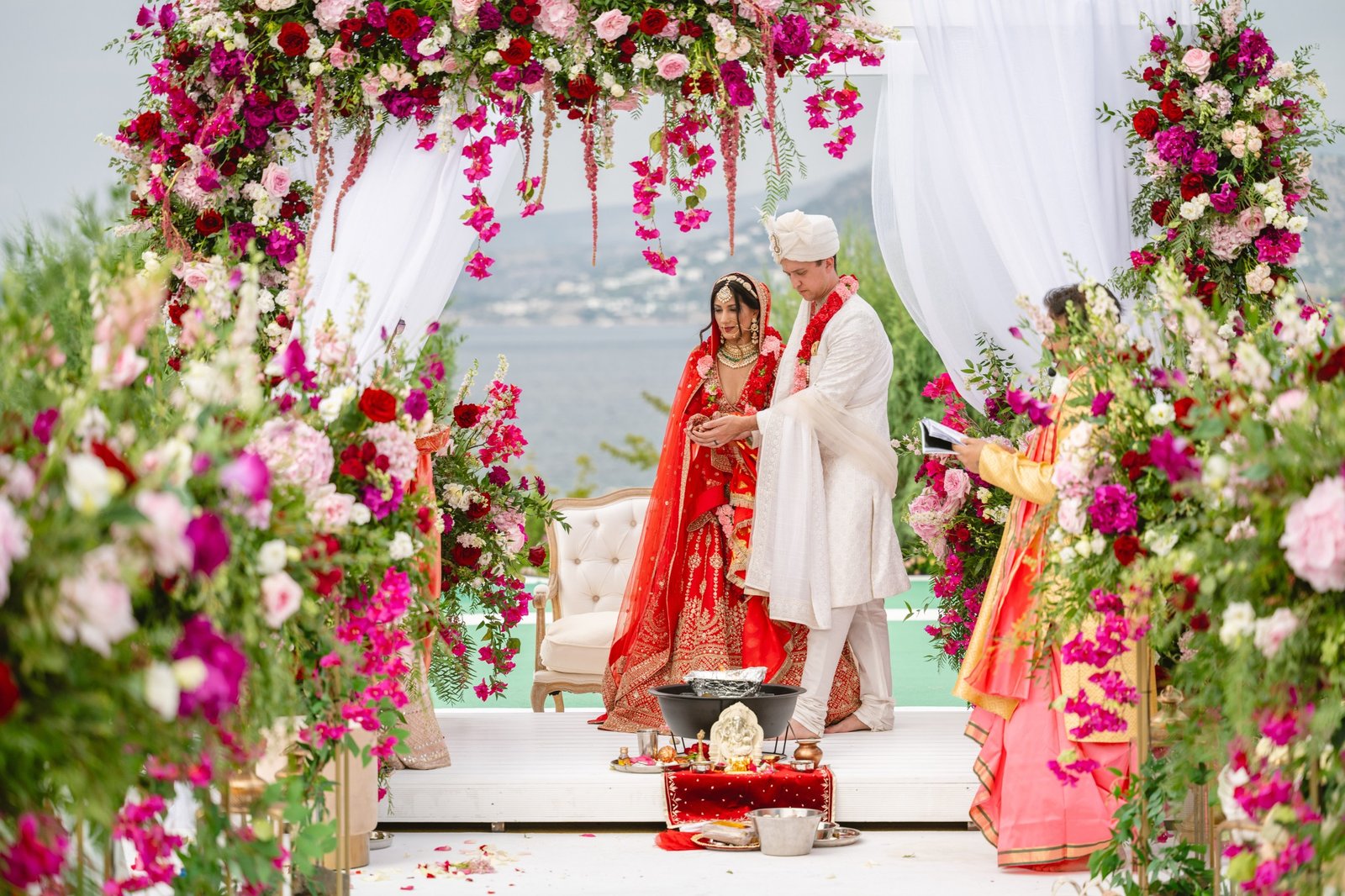 Traditional red, pink and white flower Mandap setup for a Hindu ceremony overlooking the Aegean Sea with Bride in red bridal Lehenga and groom in a white Sherwani performing a ritual 