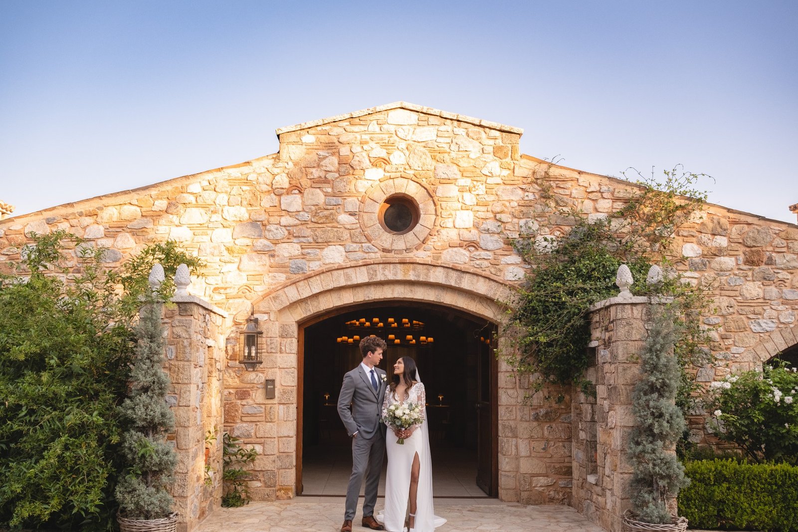 Bride in a white wedding dress next to the groom in a gray suit with Pyrgos Petreza wedding venue manor in the background