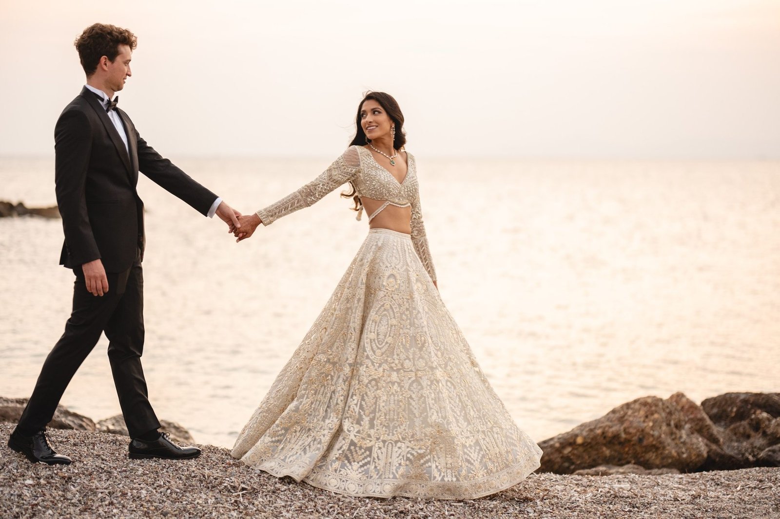 Bride wearing a white gold foil Falguni Shane Peacock Lehenga and Groom in a black tuxedo walking holding hands and smiling and walking on the shore in Athenian Rivera 