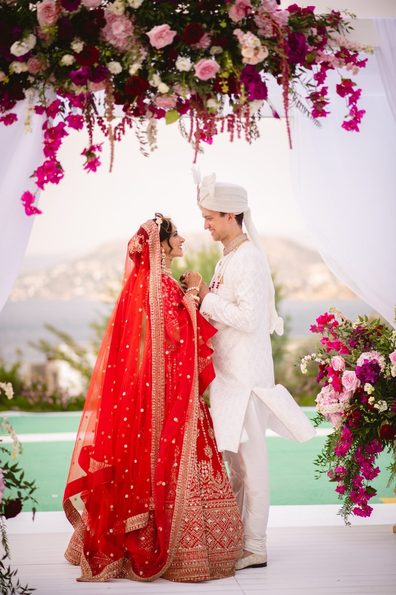 Bride in red bridal Lehenga and groom in a white Sherwani holding hands and smiling at an outdoor destination Indian wedding in Greece under their Mandap setup for a Hindu ceremony overlooking the Aegean Sea