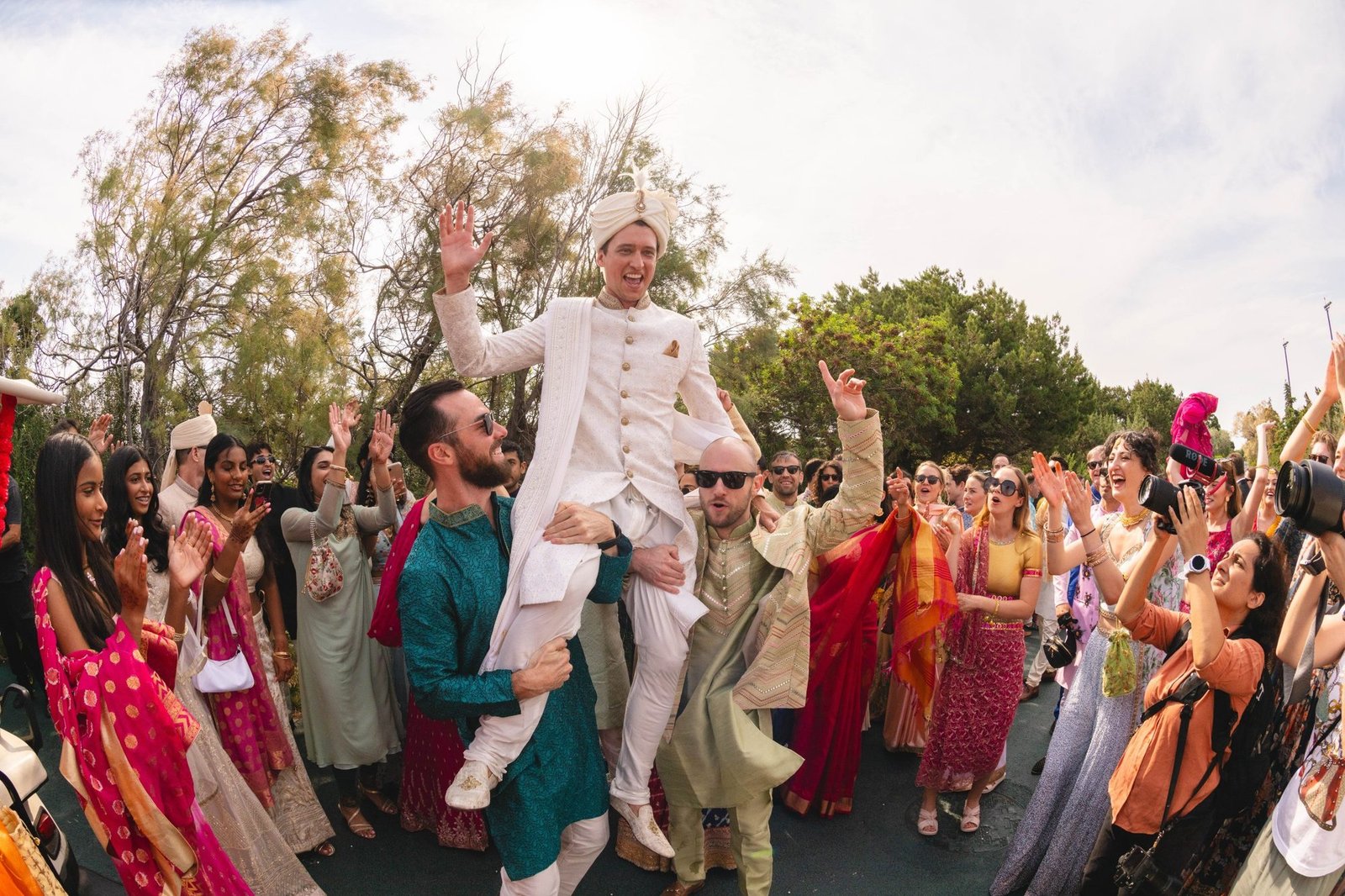 Group picture of a groom in white Sherwani being lifted by his guests celebrating his Baraat at the Grand Resort Lagonissi helipad