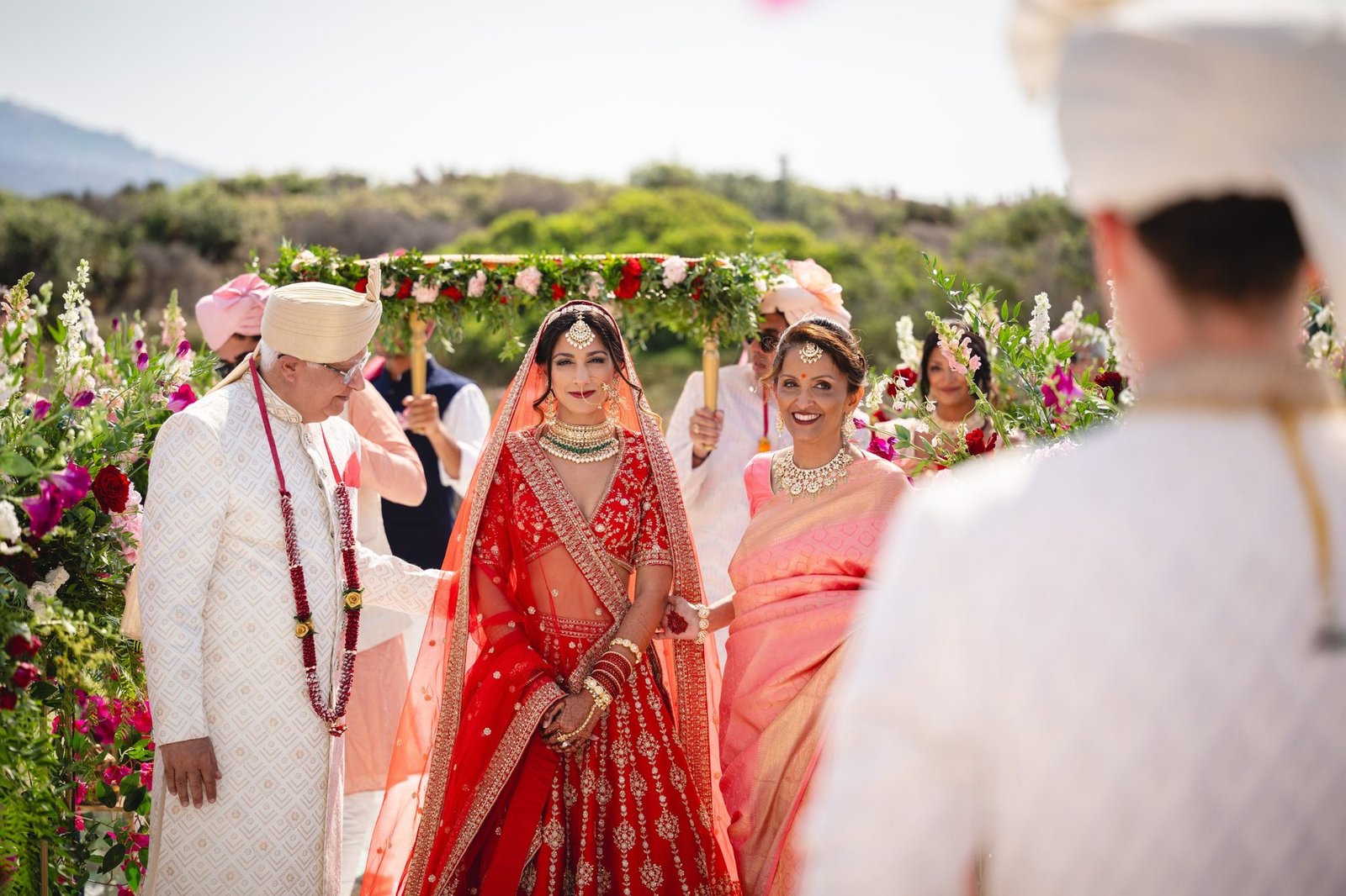 Bride in red bridal Lehenga arriving with her parents to the ceremony seeing her groom in a white Sherwani at an outdoor destination Indian wedding in Greece for the first time