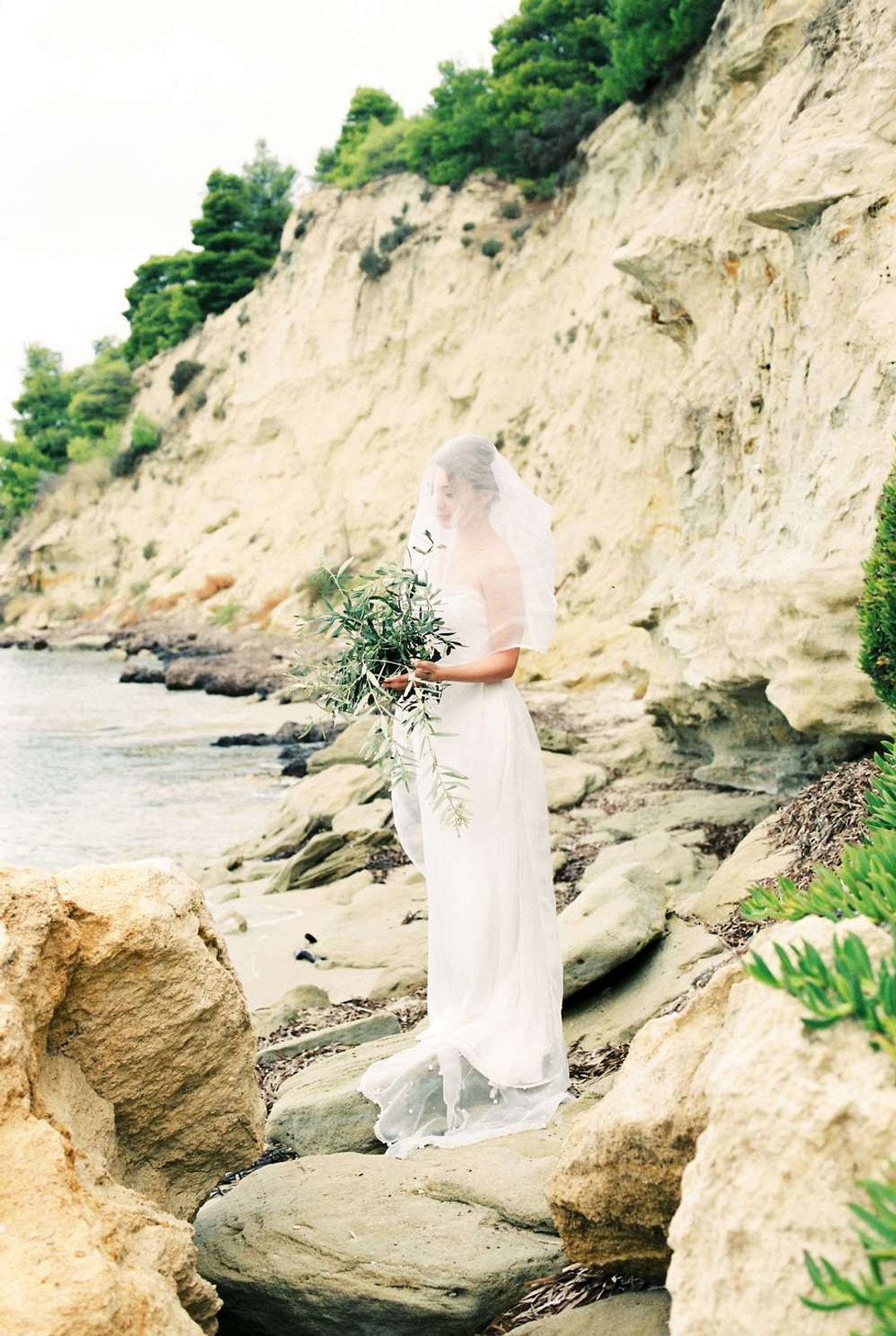 Bride waring white veil with bouquet of olive branches posing on the rock with a cliff behind her on the beach in Greece at her intimate wedding
