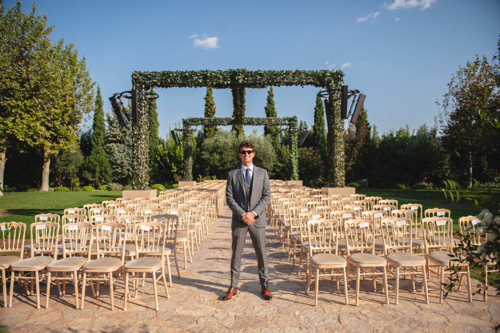Groom in grey suit standing and smiling for a portrait between rows of chairs at their Christian wedding ceremony at Pyrgos Petreza  in Athens, Greece