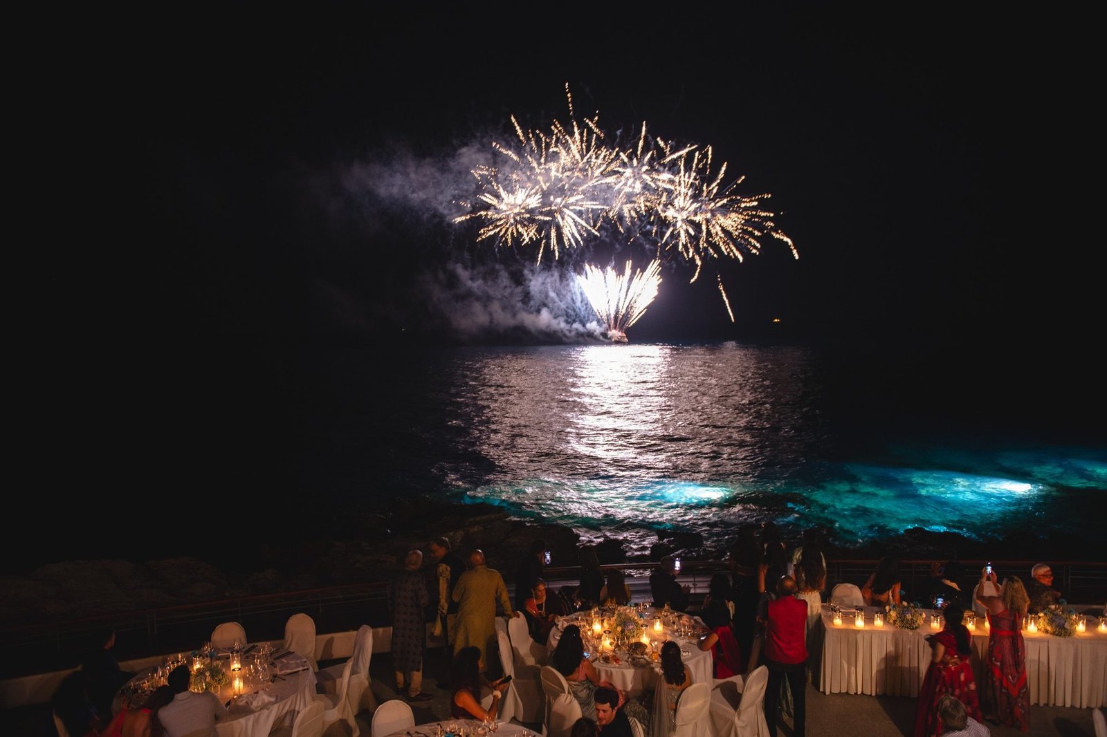 Fireworks on the water at outdoor Sangeet night event with fairy lights overlooking the sea at Veghera Lounge in Greece