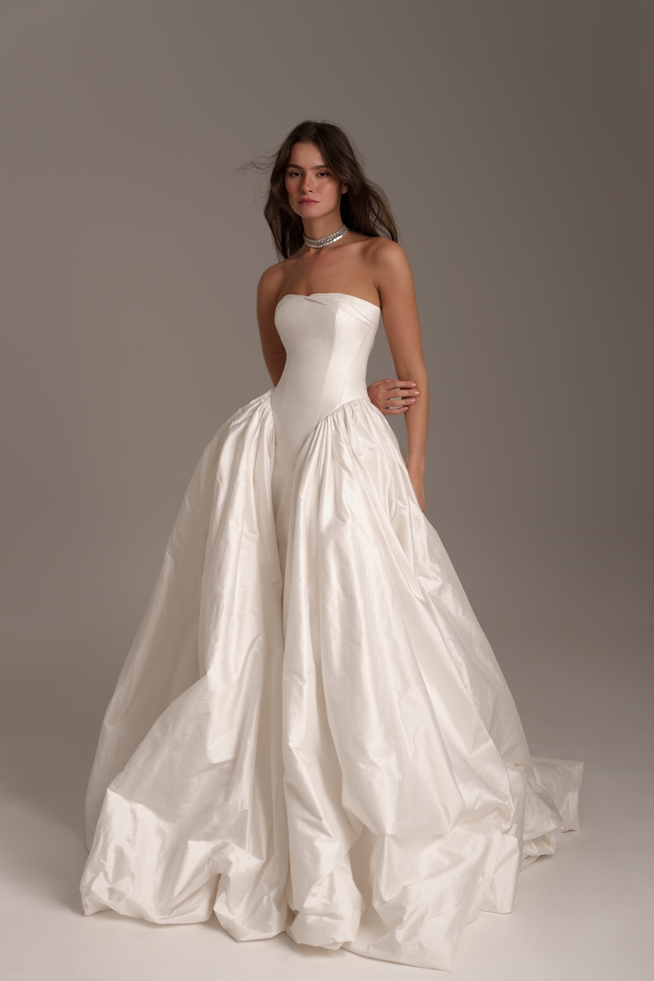 Nelly by Lihi Hod is a dramatic silk Shantung ball gown featuring a strapless sweetheart bodice and a flattering dropped waist. The structured corset flows into a voluminous skirt with rich architectural folds, a low back, and a striking cathedral-length train.