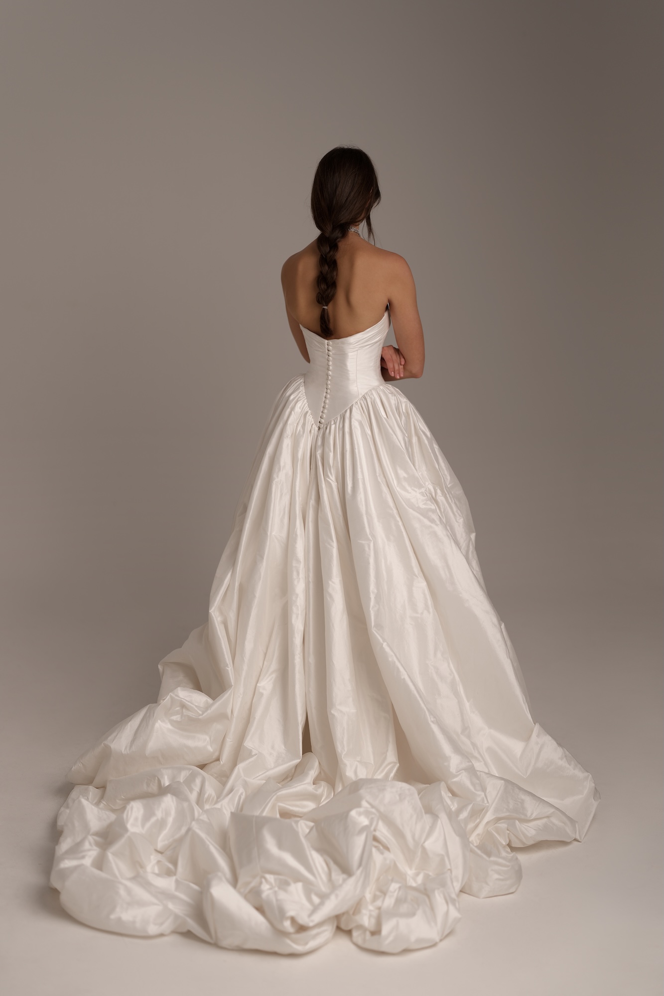 Back of Nelly, a dramatic silk Shantung ball gown featuring a strapless sweetheart bodice and a flattering dropped waist. The structured corset flows into a voluminous skirt with rich architectural folds, a low back, with fabric-covered buttons and a striking cathedral-length train.