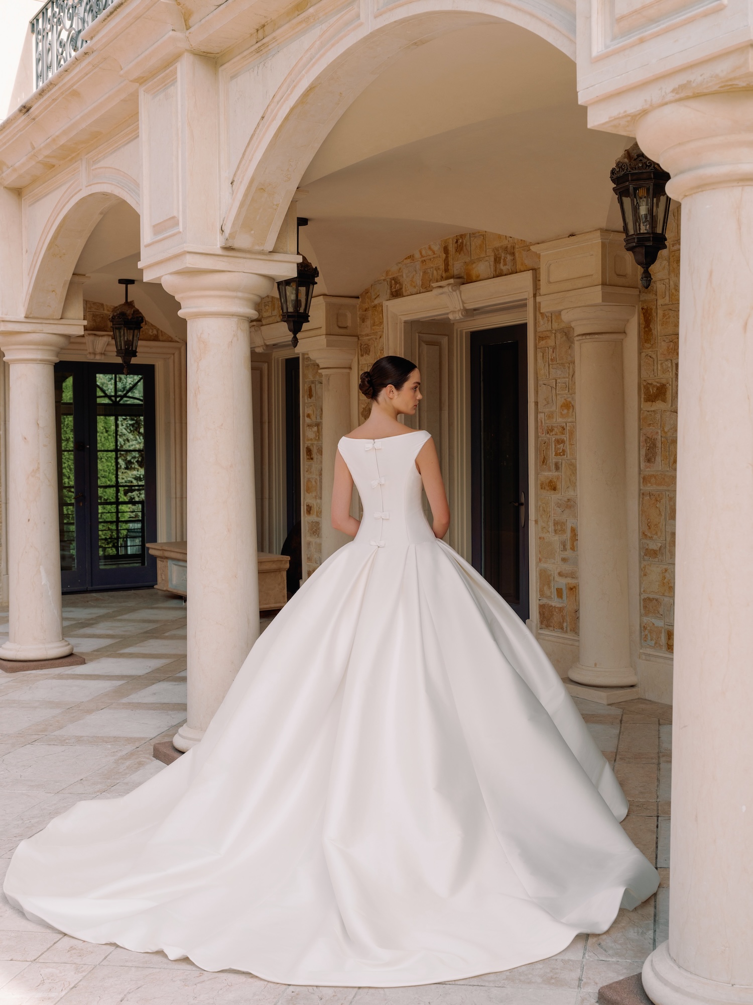 Bride standing under the Greek style church with arches in a luxury wedding dress with Italian stretch mikado fabric, with straight waist, bow detail up the center in the back, and gathered ballgown skirt.