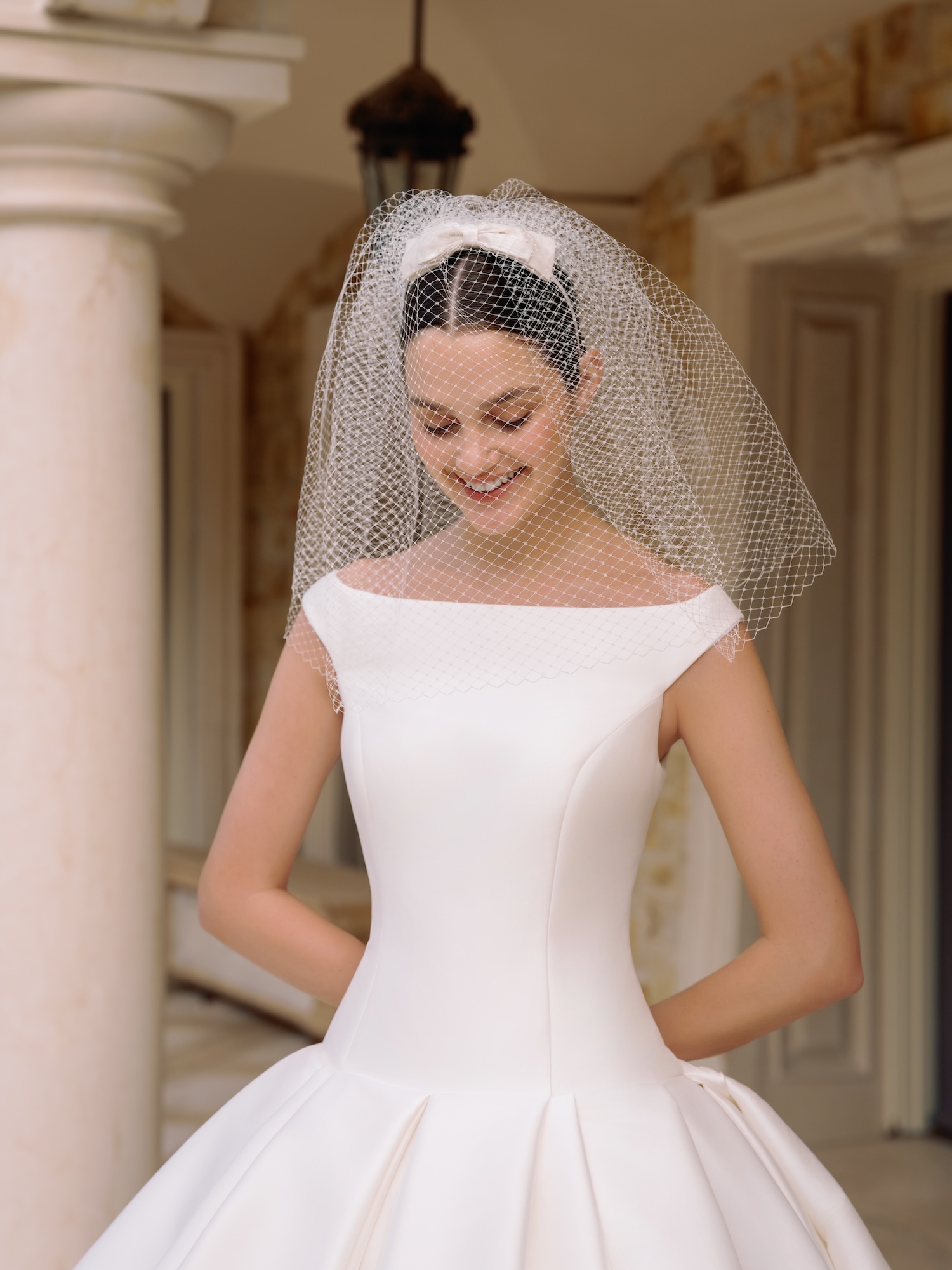 Portrait of a bride with veil over her face smiling under the Greek style church with arches in a luxury wedding dress with boat neckline,  straight waist, and gathered ballgown skirt.
