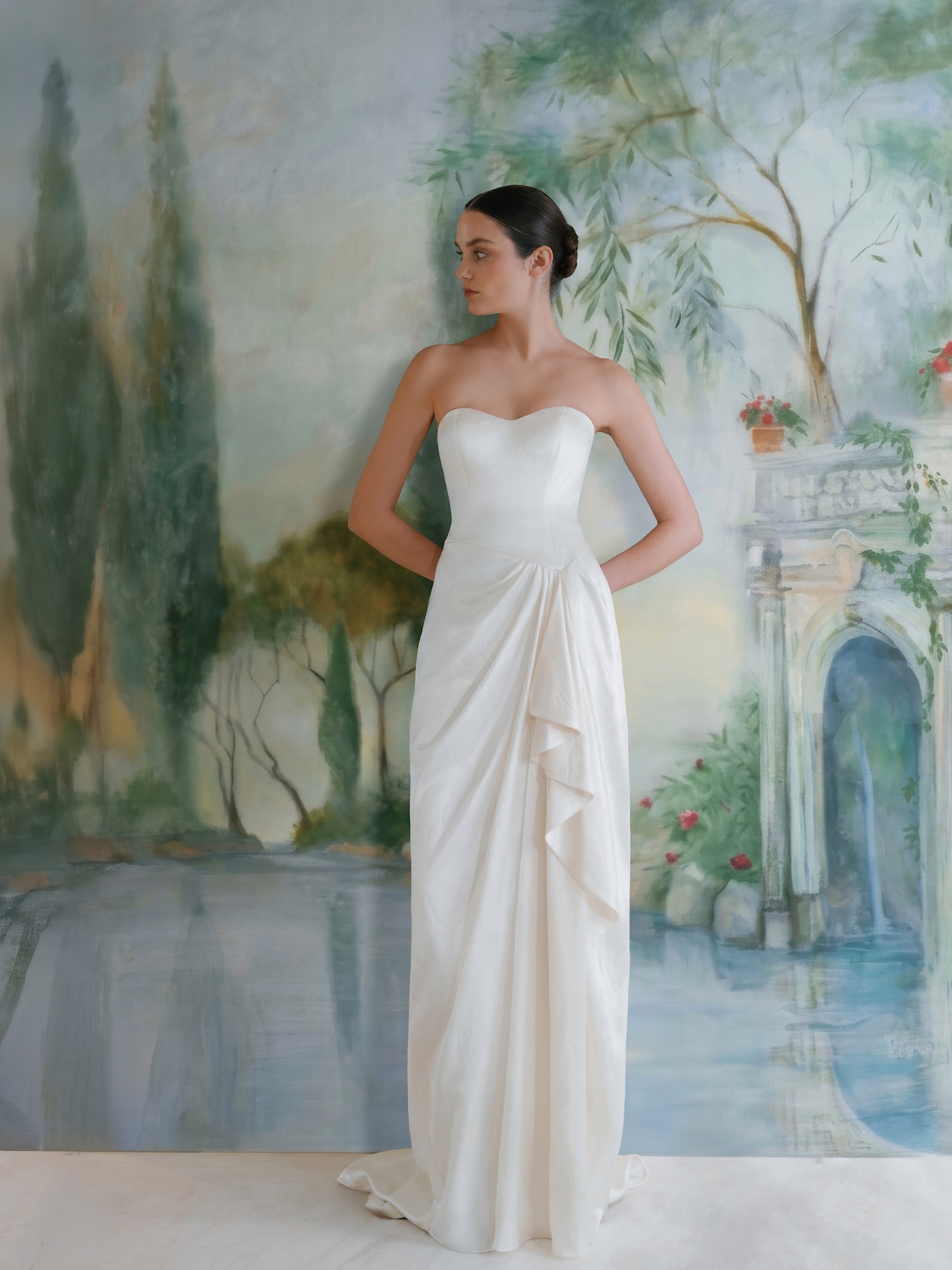 Bride in embossed charmeuse fabric wedding dress with soft sweetheart neckline, asymmetrical drop waist bodice and gathered ruffle with draping in the column skirt.
