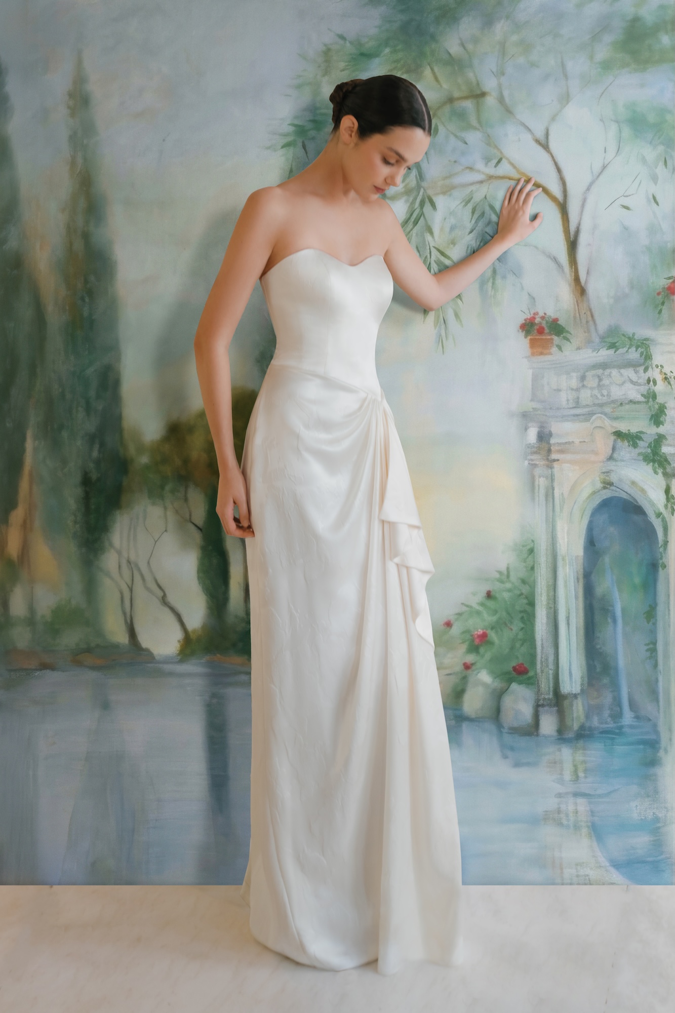 Bride in front of the fine art wall fresco in Greek style wedding dress with soft sweetheart neckline, asymmetrical drop waist bodice and gathered ruffle with draping in the column skirt.