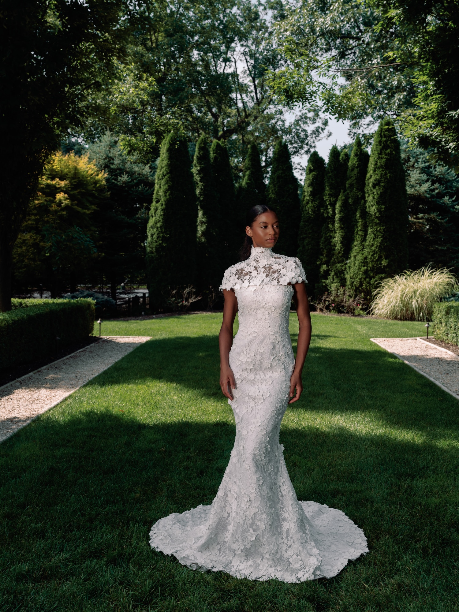 Bride standing in the luxury  green garden in timeless fitted wedding dress with three dimensional embroidered lace, strapless straight across scallop trim neckline with a matching capelet on shoulders.