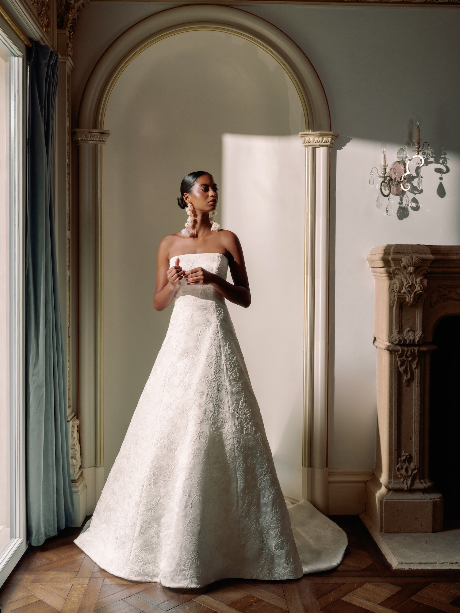 Bride with long earrings standing in a luxury room in embroidered satin wedding dress, with straight across neckline, princess seams, and modified aline skirt.