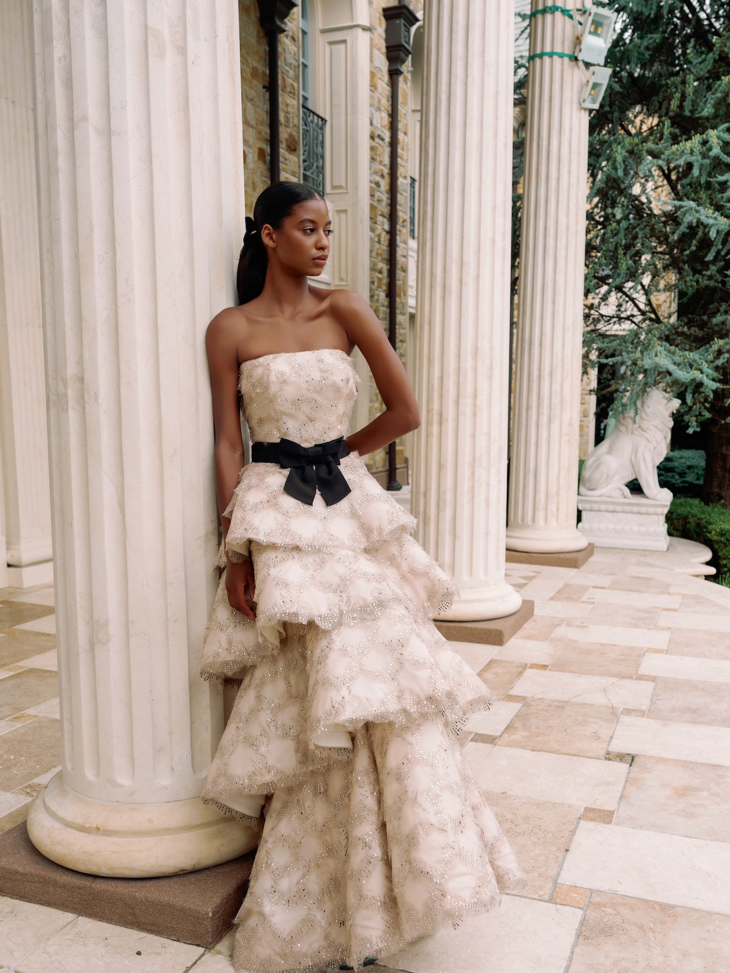 Model wearing hand beaded organza straight across neckline natural waist with tiered modified aline skirt posing with Greek columns