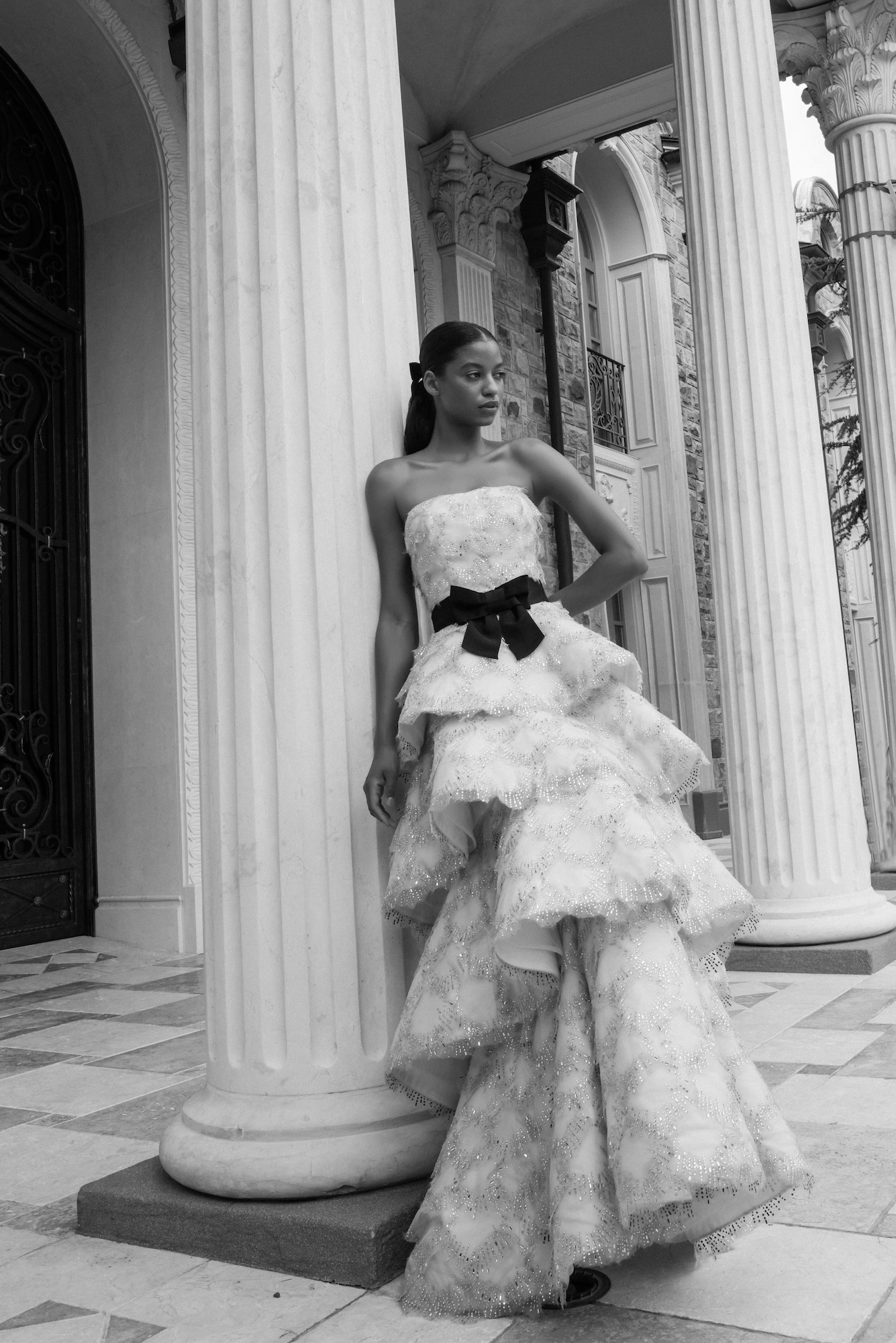 Black and white image with a model wearing hand beaded organza wedding dress with tiered modified aline skirt and black belt posing with Greek columns