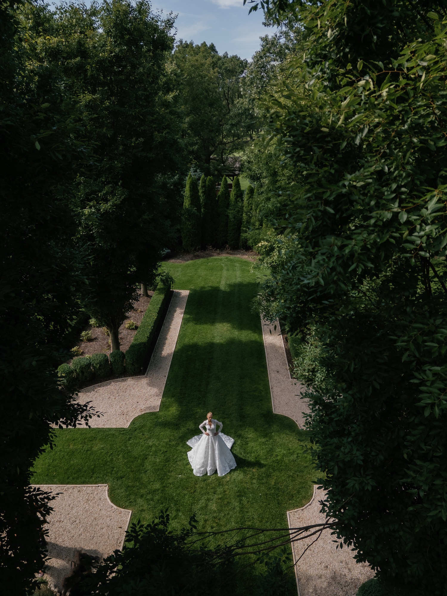 Picture from above showing a bride walking in the green garden in timeless Alencon lace wedding dress with cathedral length veil.
