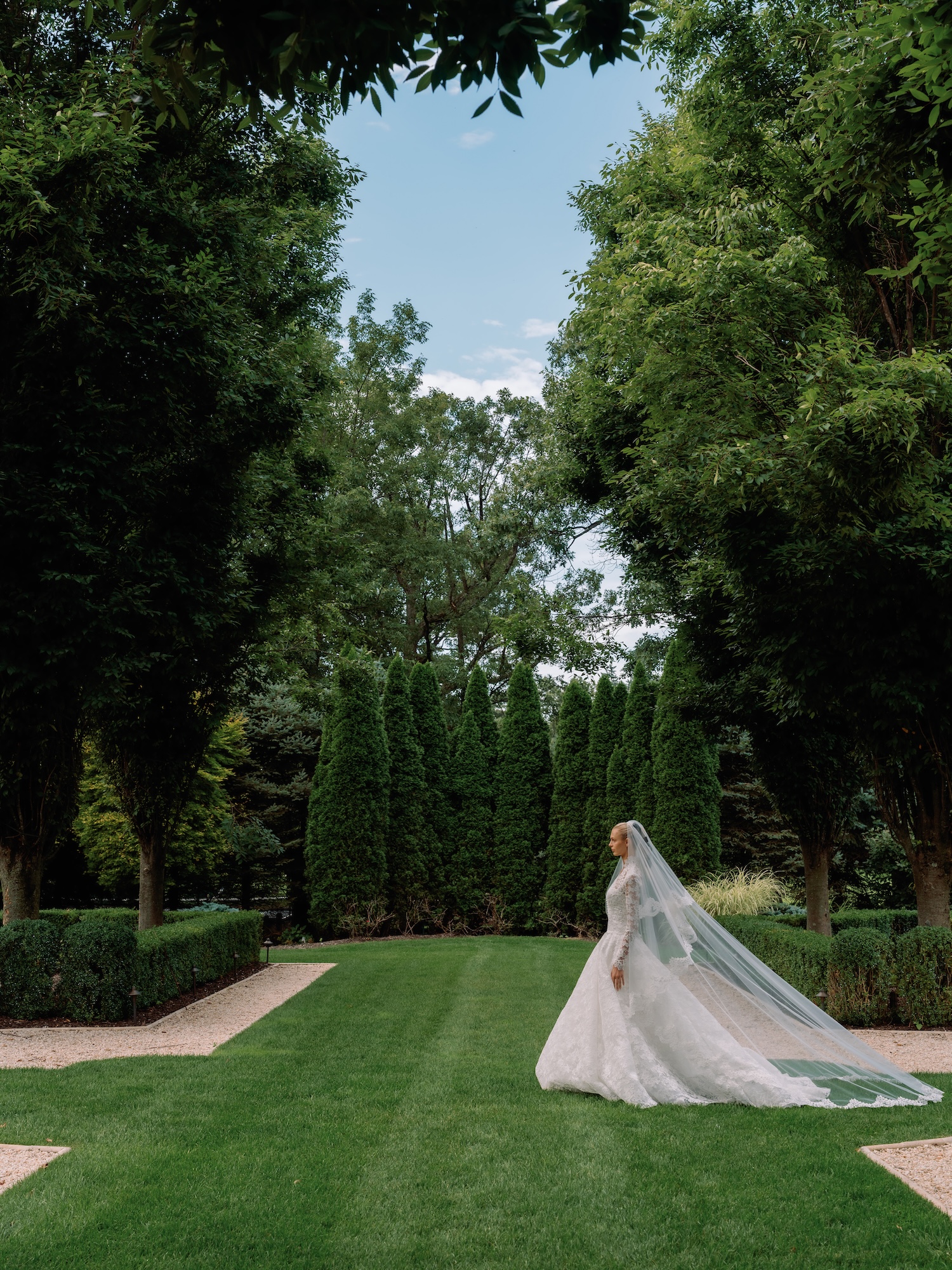 Bride walking in the green garden in timeless Alencon lace wedding dress with scalloped edge cathedral length veil with blusher including the scalloped edge detail.